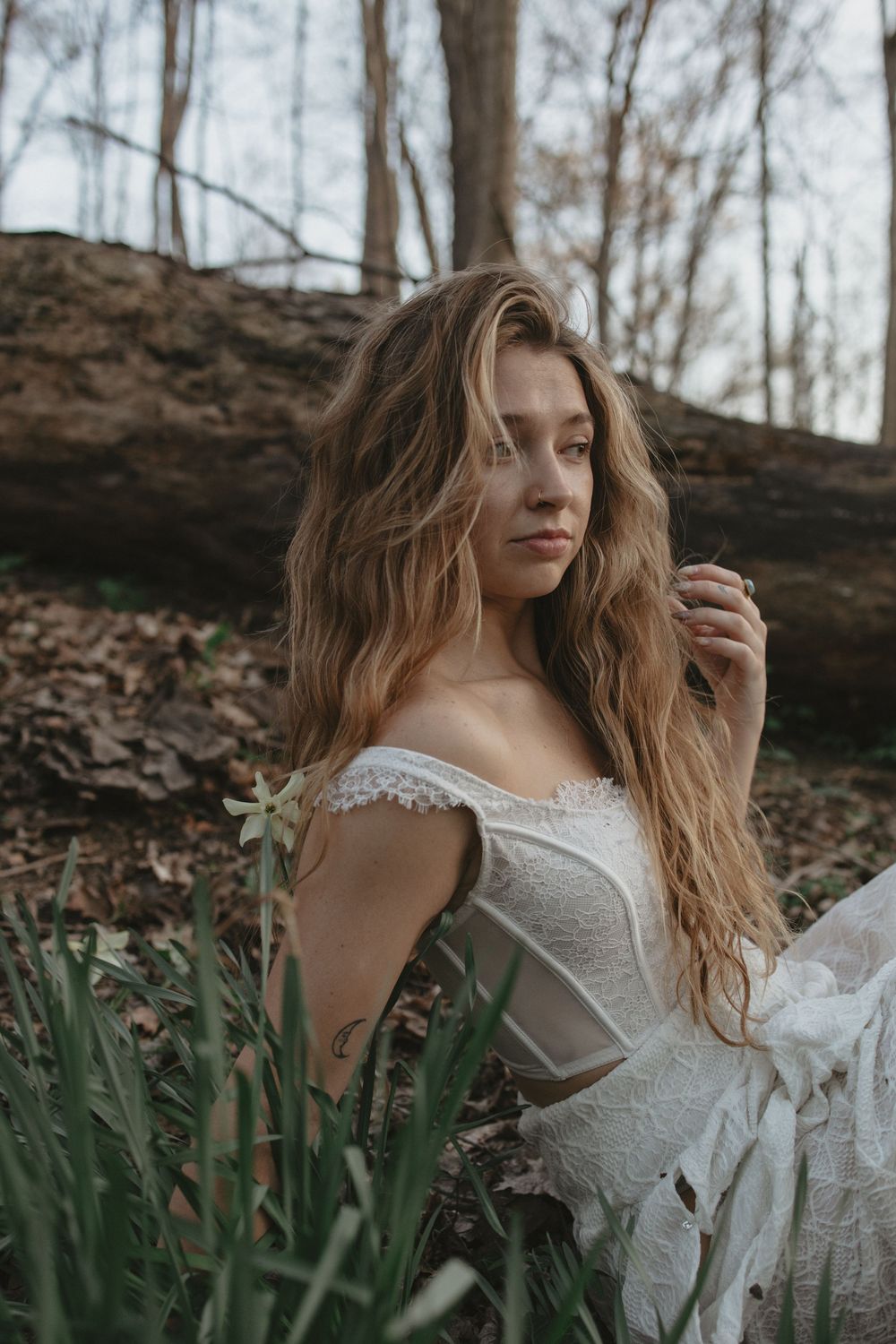 Young woman with wavy blonde hair in white lace off-shoulder dress posing in a forest setting.