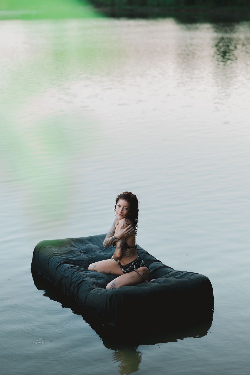 A person relaxes on a black inflatable mattress floating peacefully on calm lake waters at dusk.
