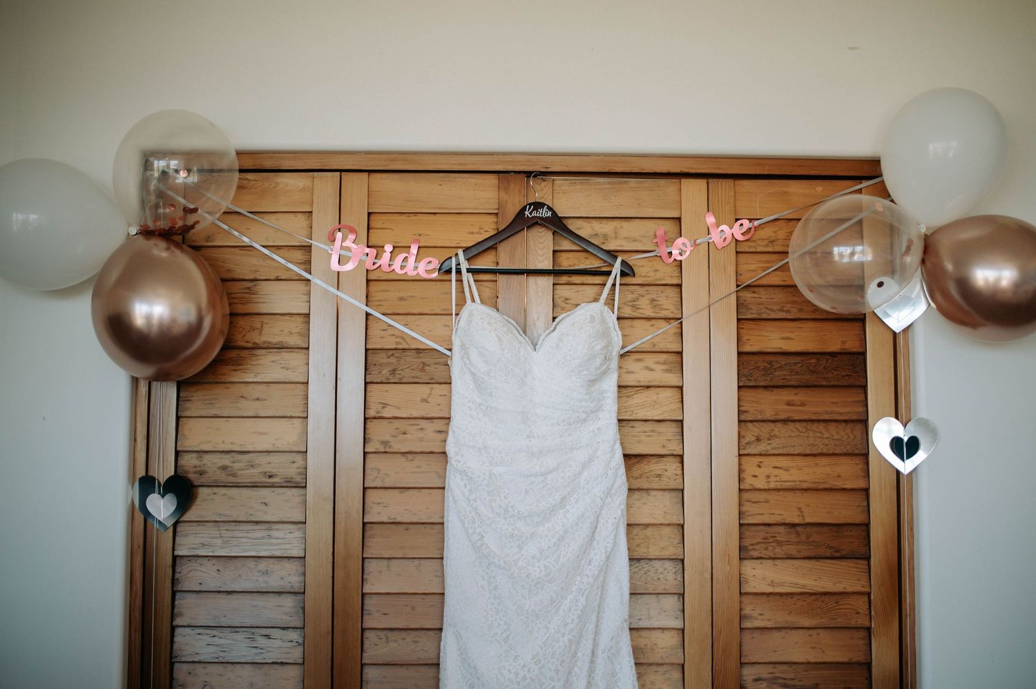 A white wedding dress hangs on display with balloons and decorations against wooden walls.