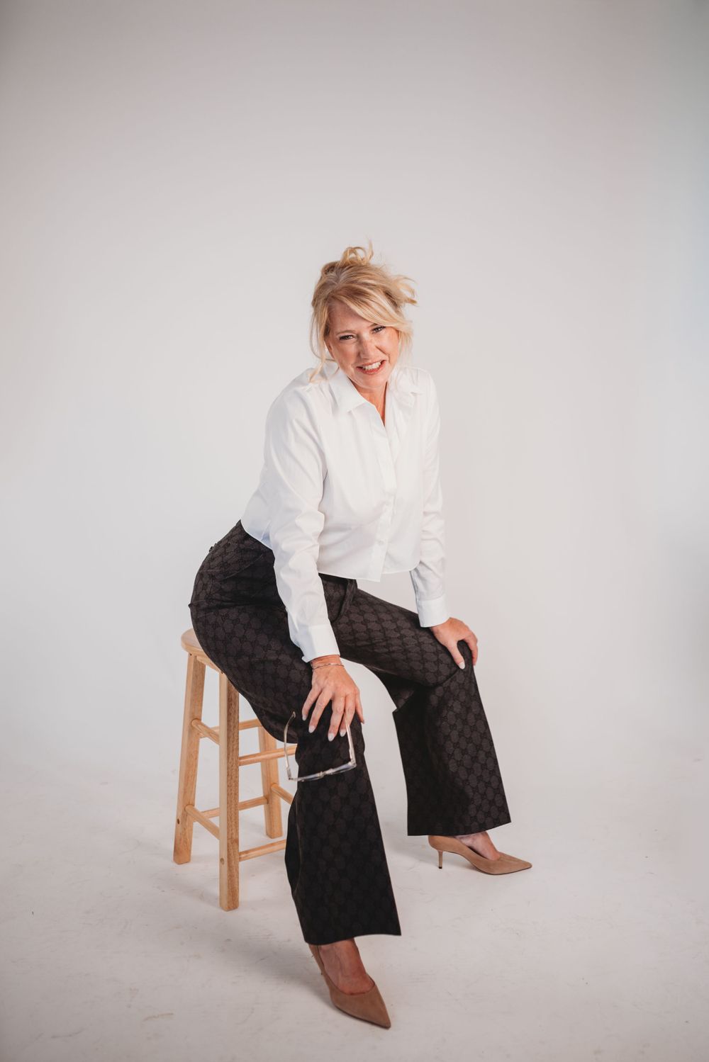 Professional portrait of someone sitting on wooden stool wearing white shirt and patterned pants with brown shoes.