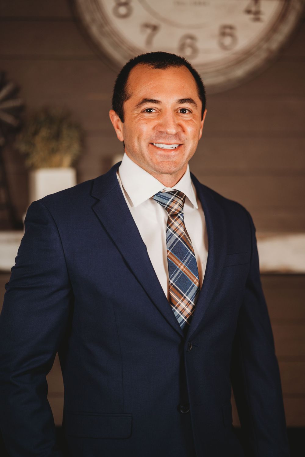 Professional headshot of person in navy suit with plaid tie against rustic wall.