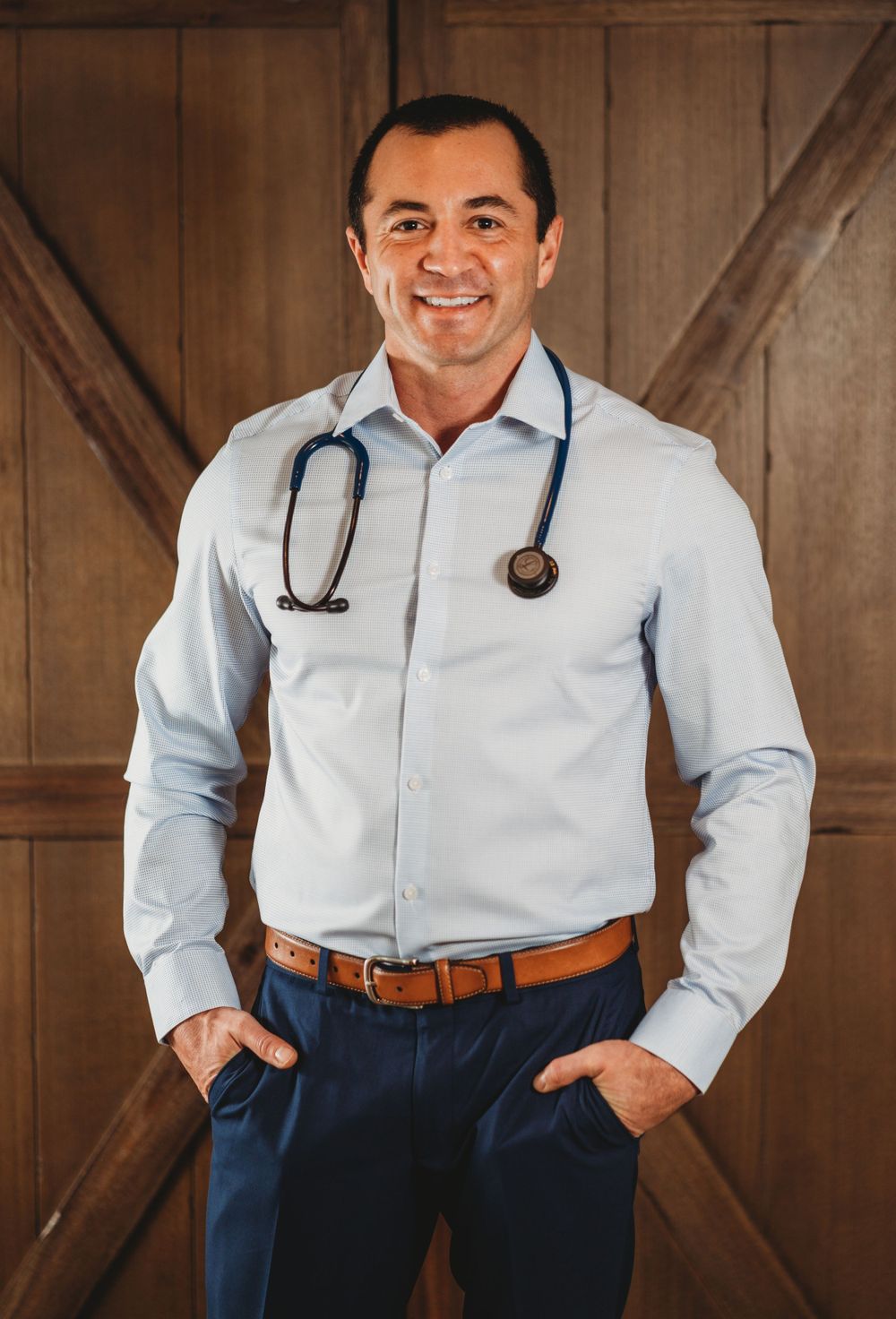 Healthcare professional in light blue shirt and navy slacks wearing stethoscope standing against wooden door background.