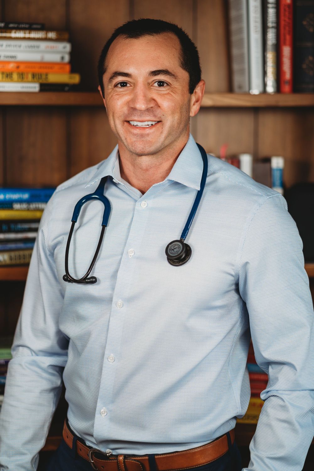 Medical professional with stethoscope and light blue dress shirt posing in front of bookshelf.
