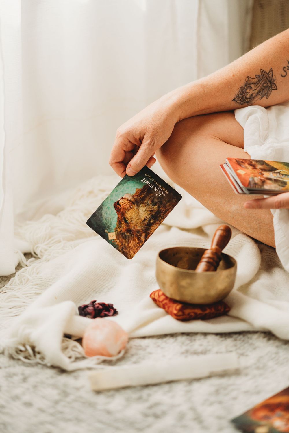 Close-up of hands holding tarot cards with singing bowl and crystals on white fabric.