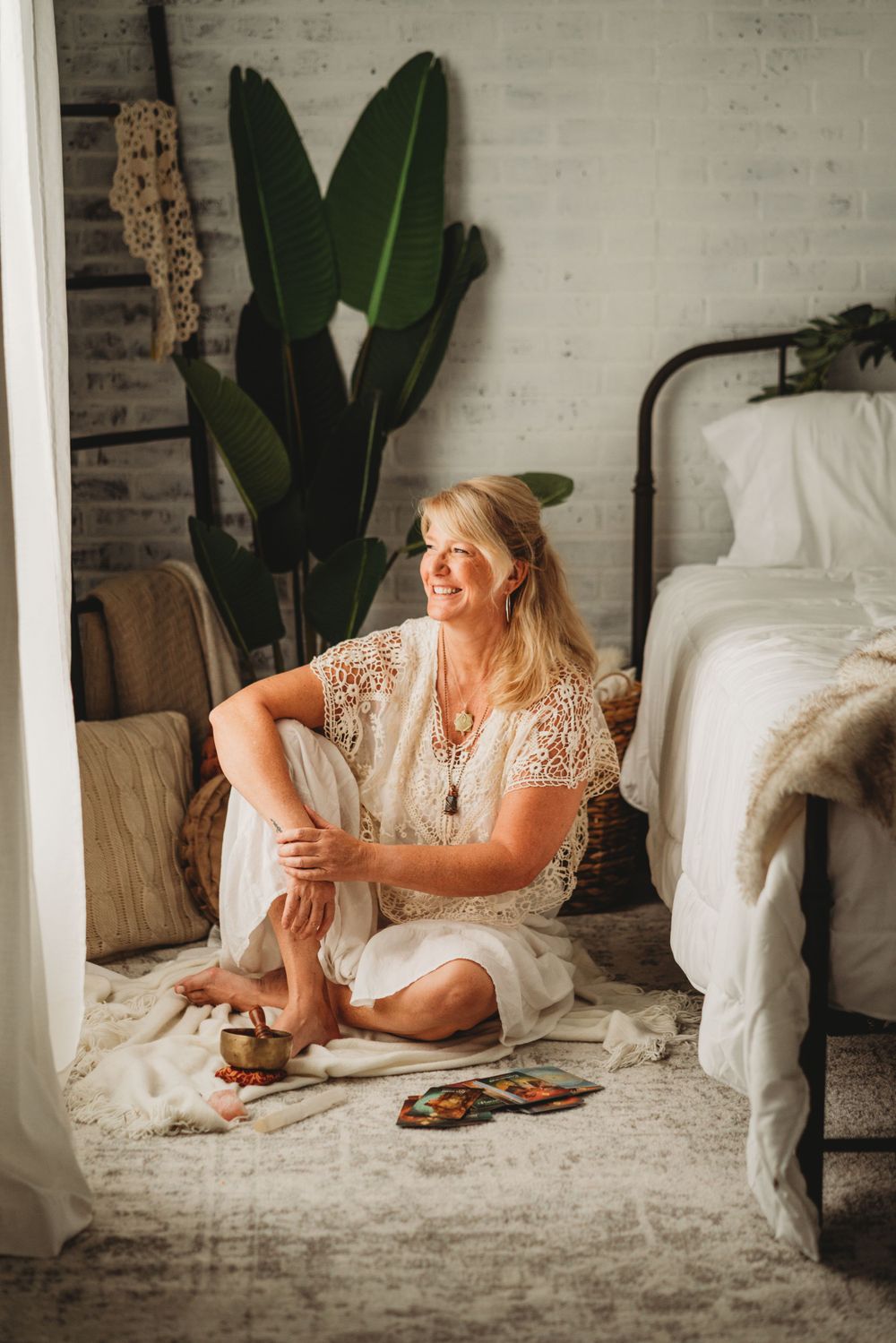 Person in white lace dress sitting on floor with cards and crystals near potted plant and bed.