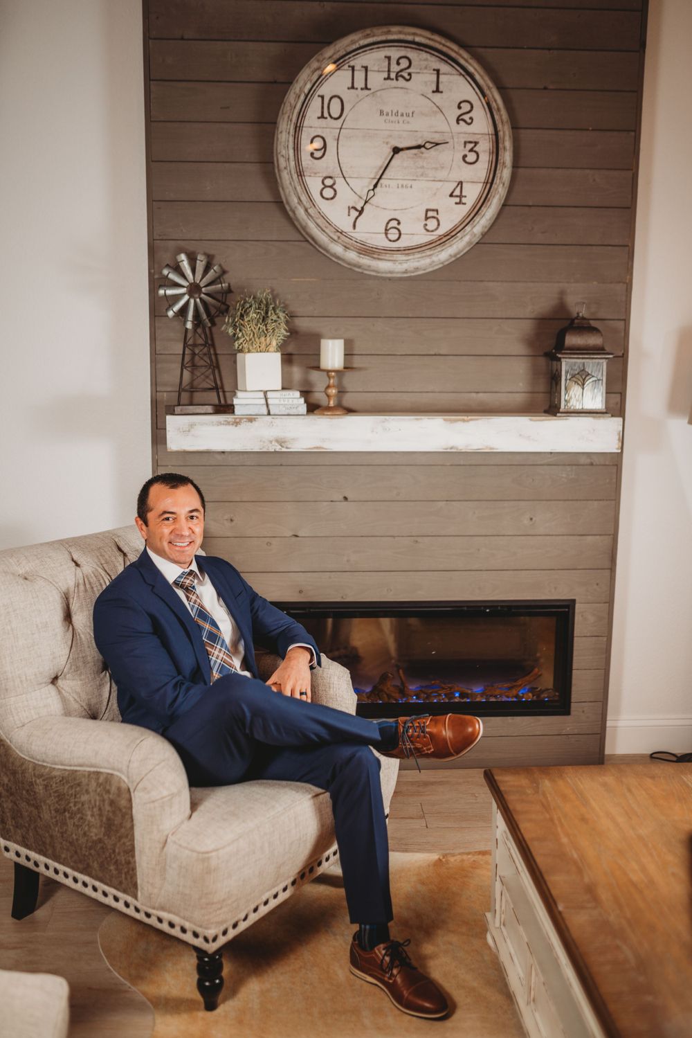 Professional in navy suit seated in beige armchair near fireplace with large clock and rustic decor.