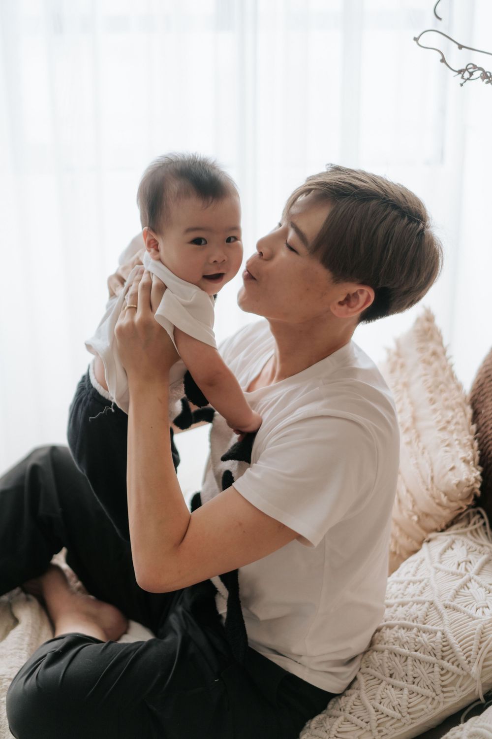 Parent and infant share a tender moment while relaxing on a cozy couch with white curtains in background.