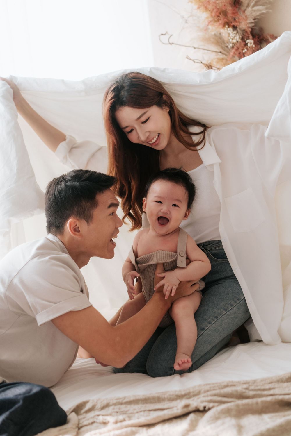 Family of three plays together under white sheets creating playful and loving atmosphere.