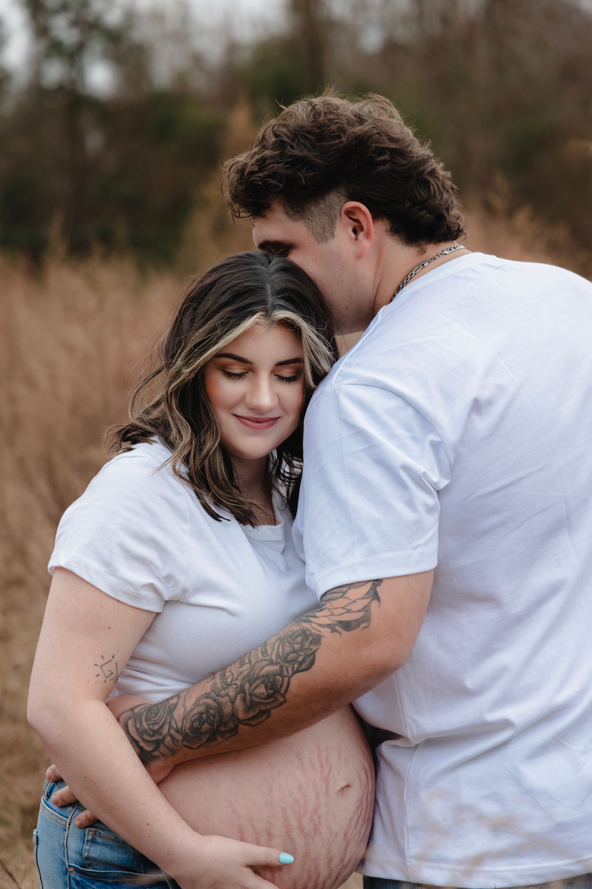 An intimate maternity photo session showing a couple in white shirts against tall grass.