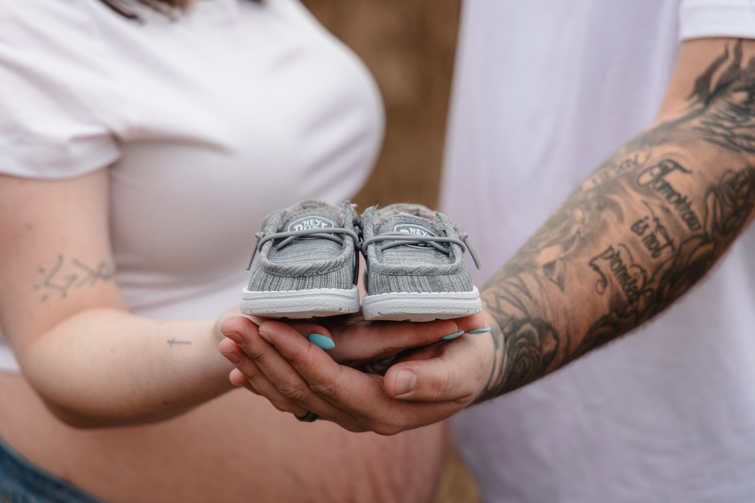 Tattooed hands hold tiny baby shoes during a pregnancy announcement photoshoot.