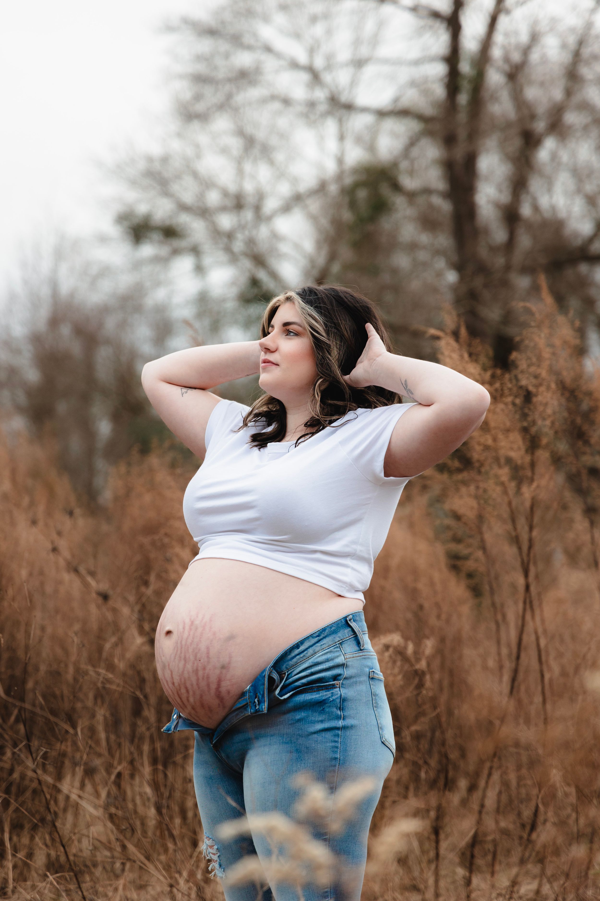 A pregnant person in white top and jeans poses naturally in a field of brown grass.