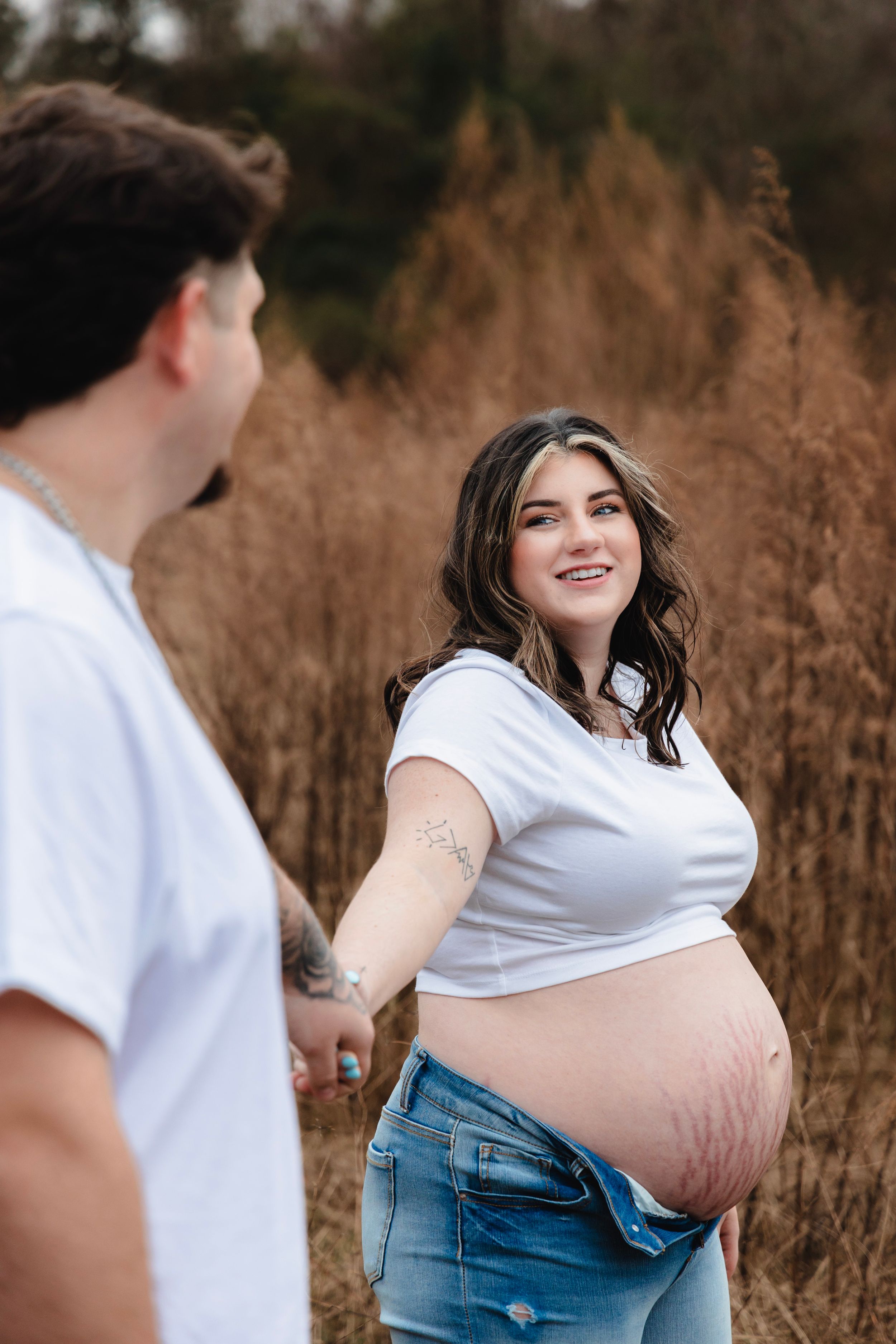 Maternity photoshoot in a field with a couple wearing white and denim showing off a baby bump.