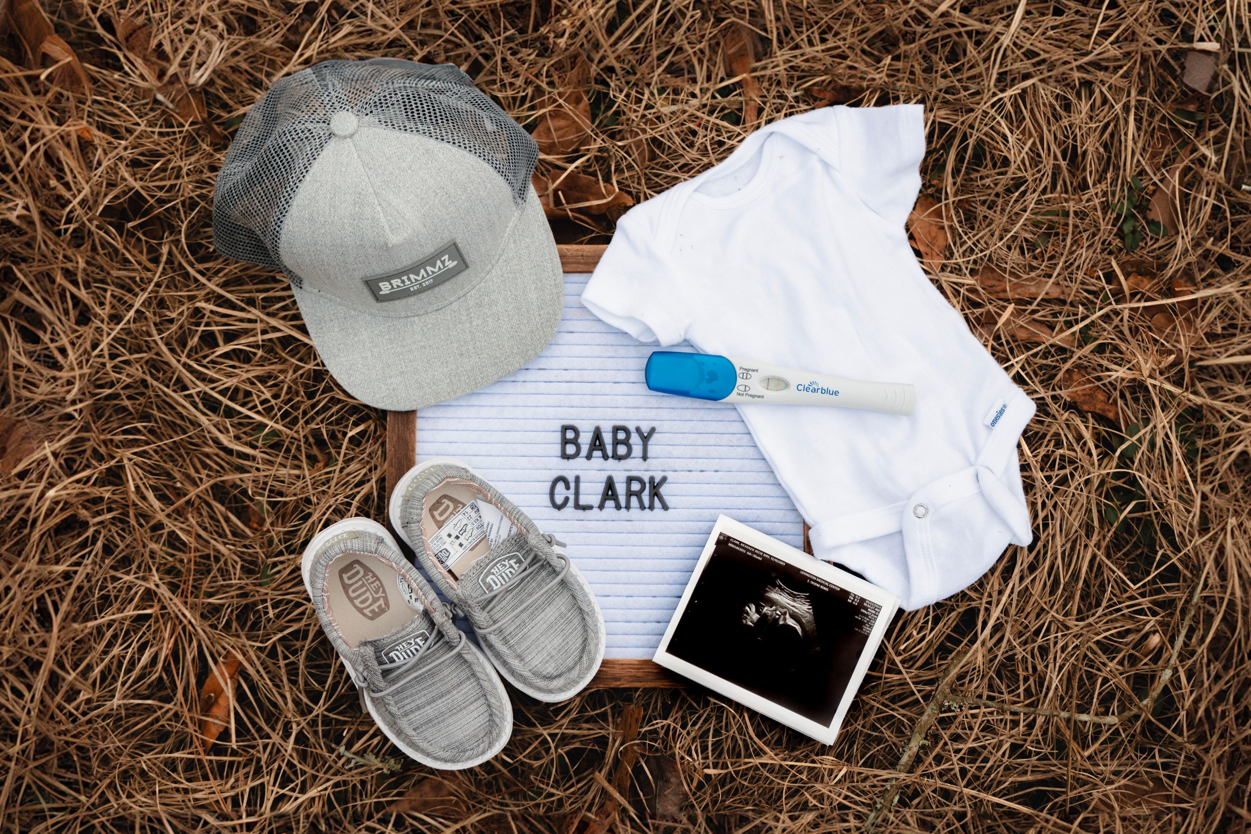 Baby items including shoes, ultrasound photo, and personalized onesie laid out on brown grass.