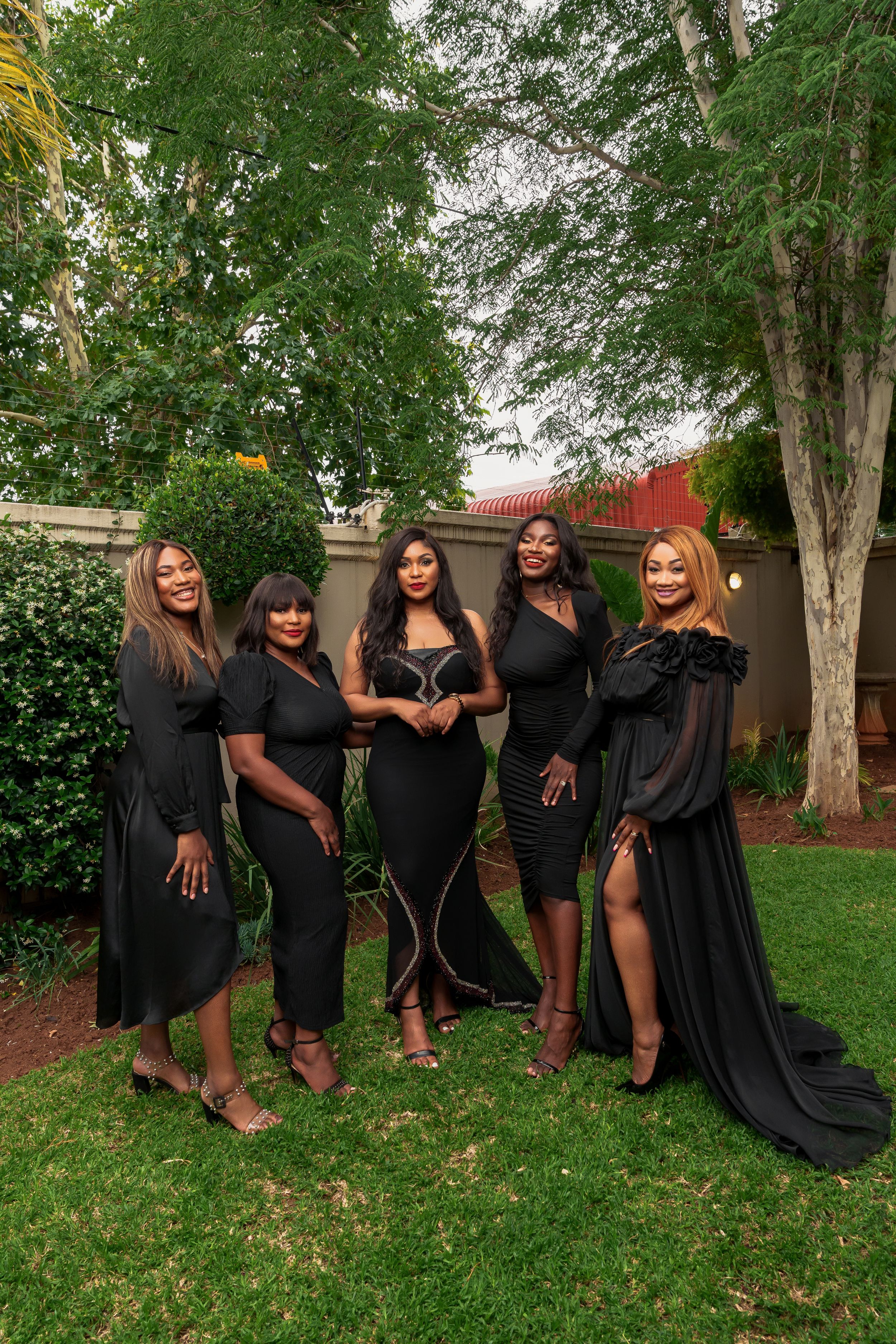 A group of five women pose together wearing elegant black dresses in a garden setting.