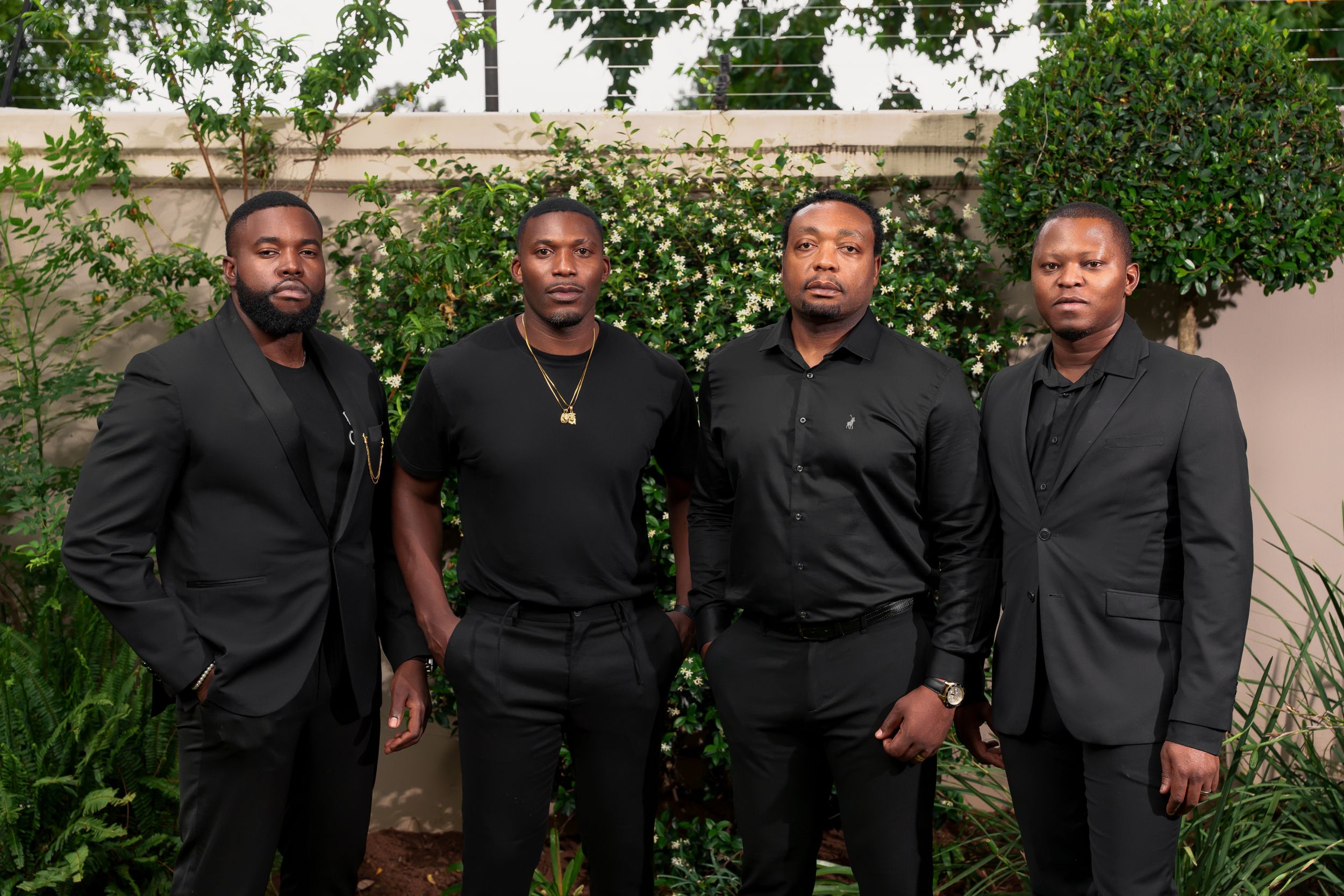 Four men in matching black formal wear pose together in outdoor garden setting.