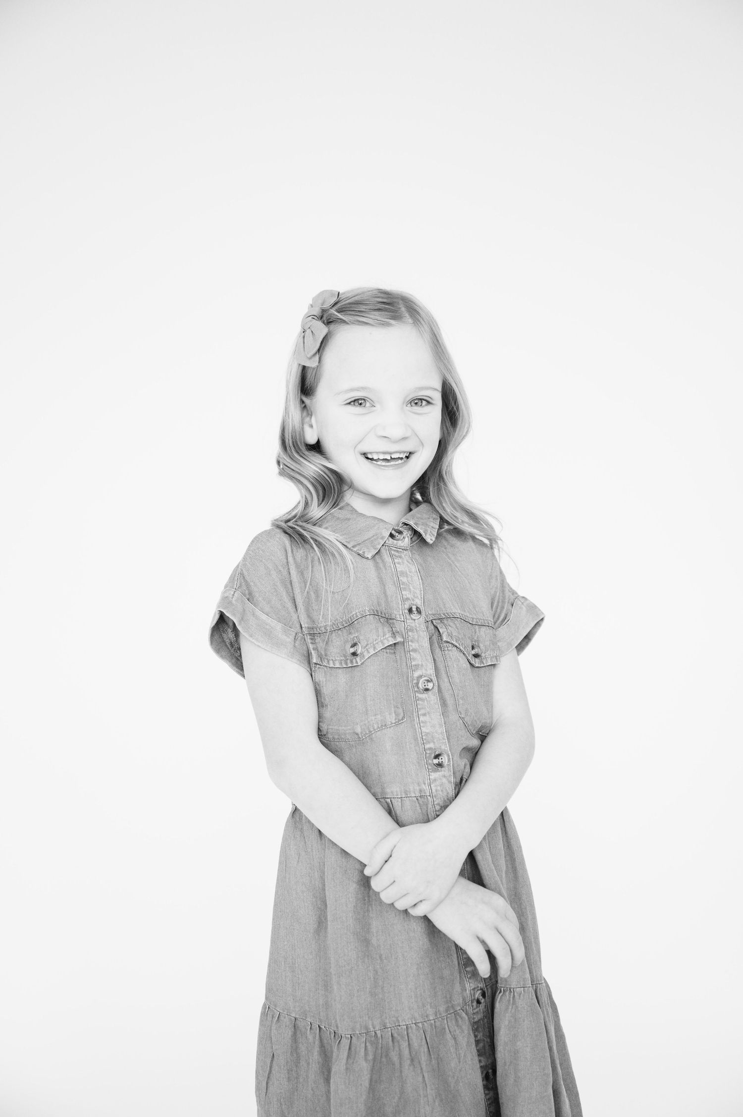 Black and white portrait of a young girl wearing a denim dress and smiling against a white backdrop.
