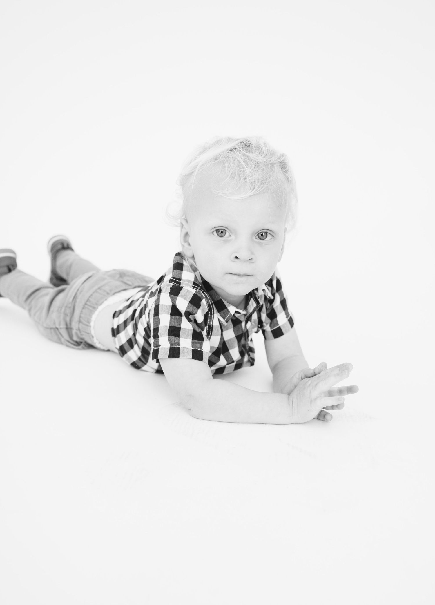Black and white photo of a toddler in checkered shirt lying on their stomach against white background.