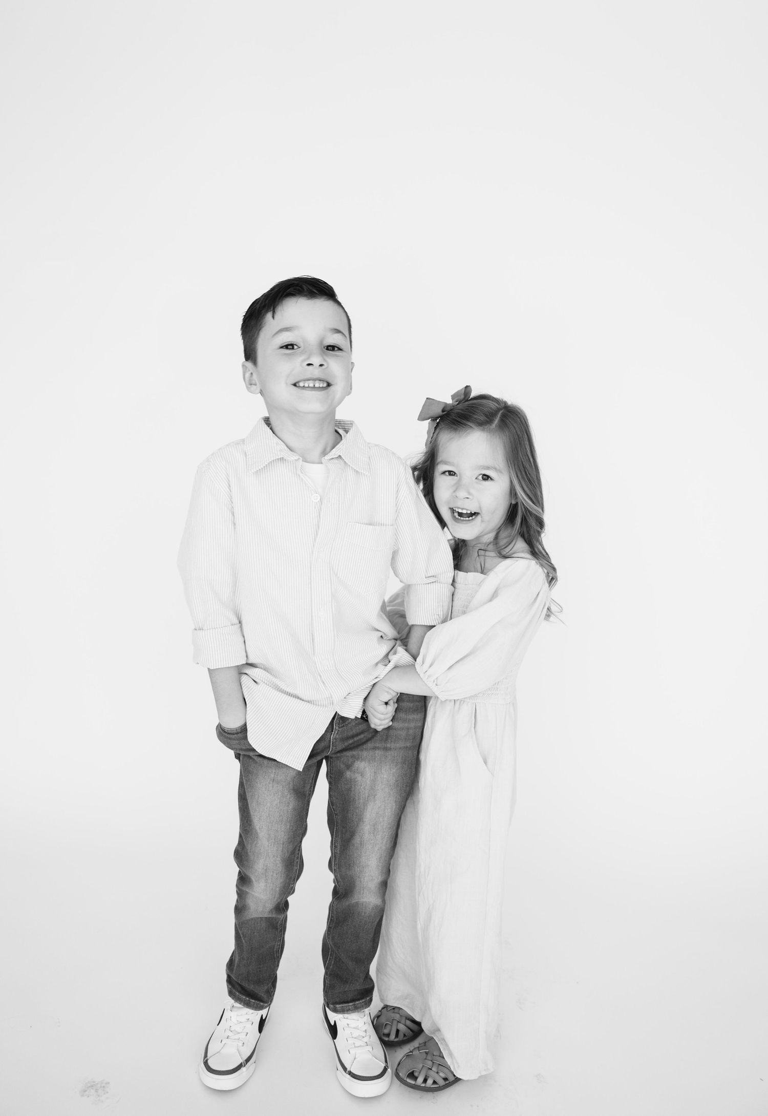Black and white photo of two children standing together, wearing casual attire against white backdrop.