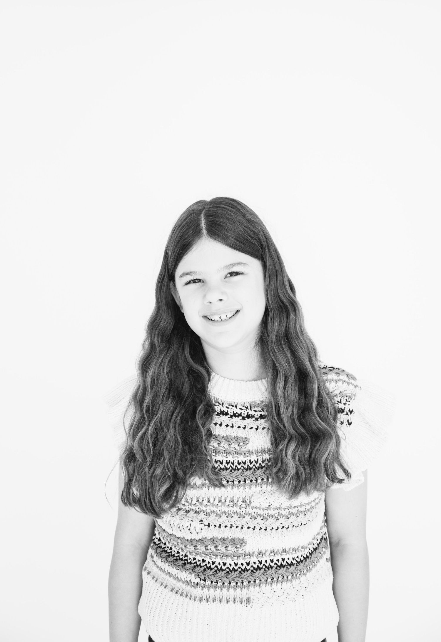 Black and white portrait of long-haired child wearing patterned top against white background.