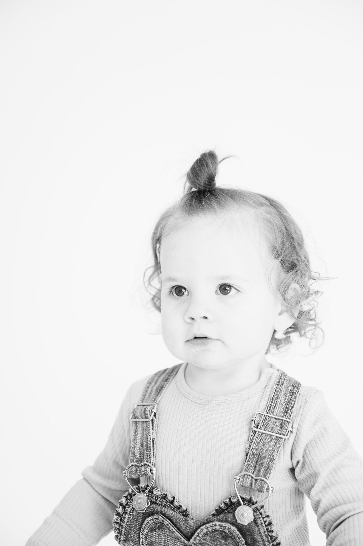 Black and white close-up portrait of toddler in overalls with top knot hairstyle.