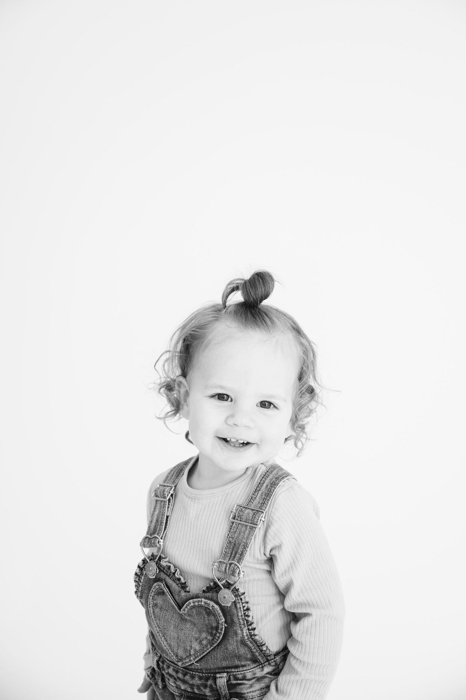 Black and white portrait of toddler in overalls with curly hair and sweet smile.