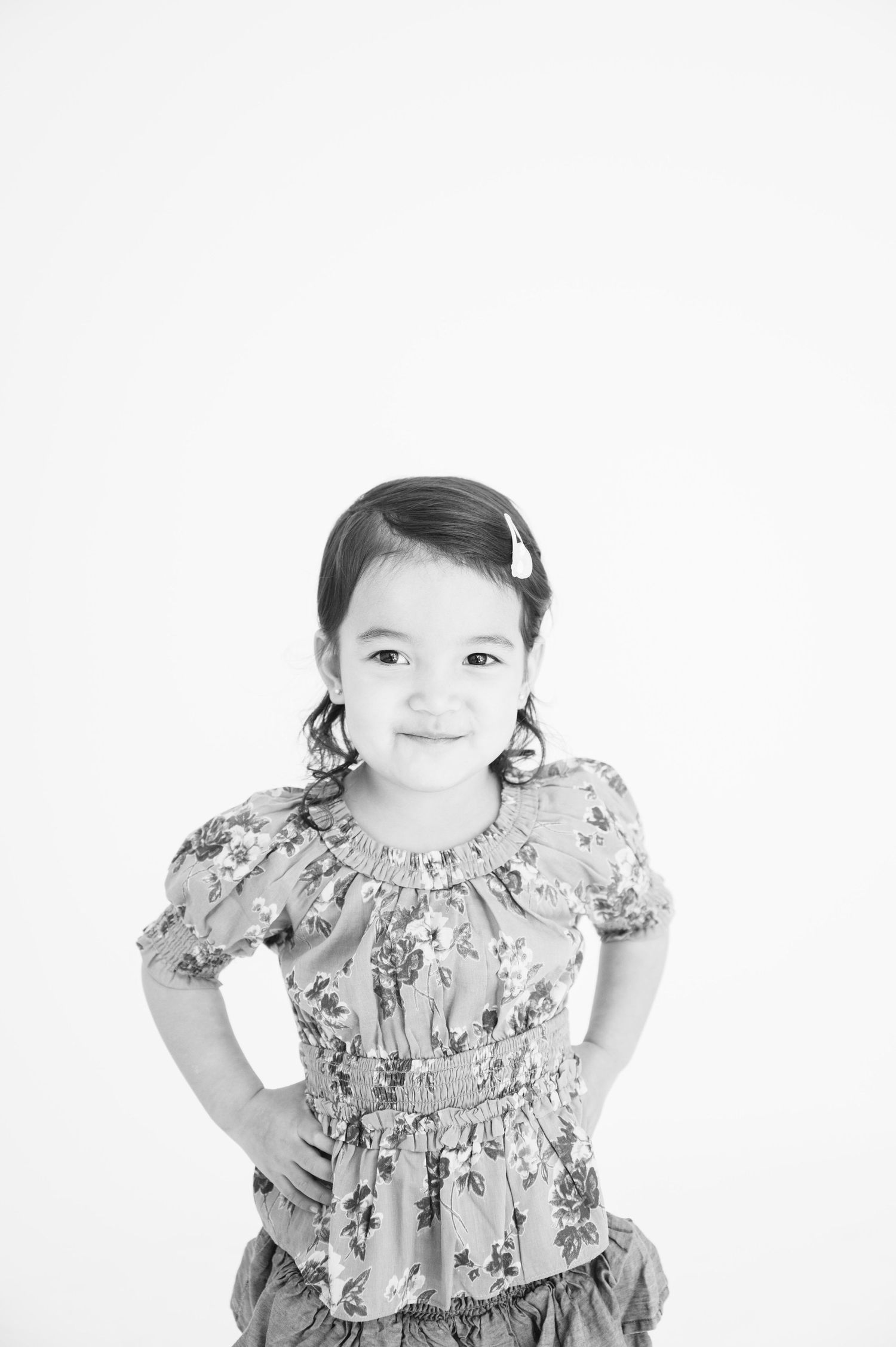 Black and white photo of a child in a floral dress with pigtails smiling against white background.