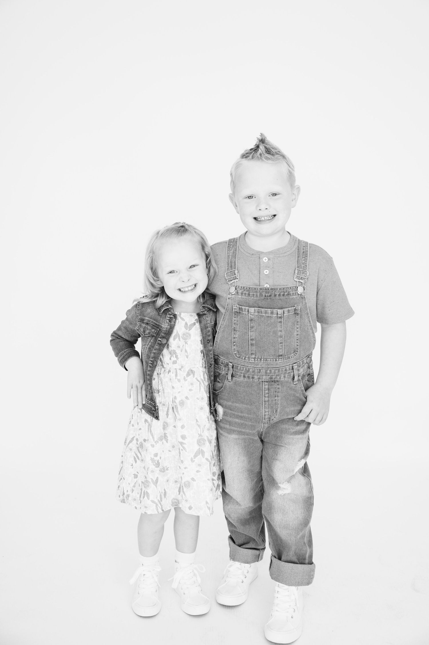 Black and white photo of two children, one in overalls and one in a dress, standing together.