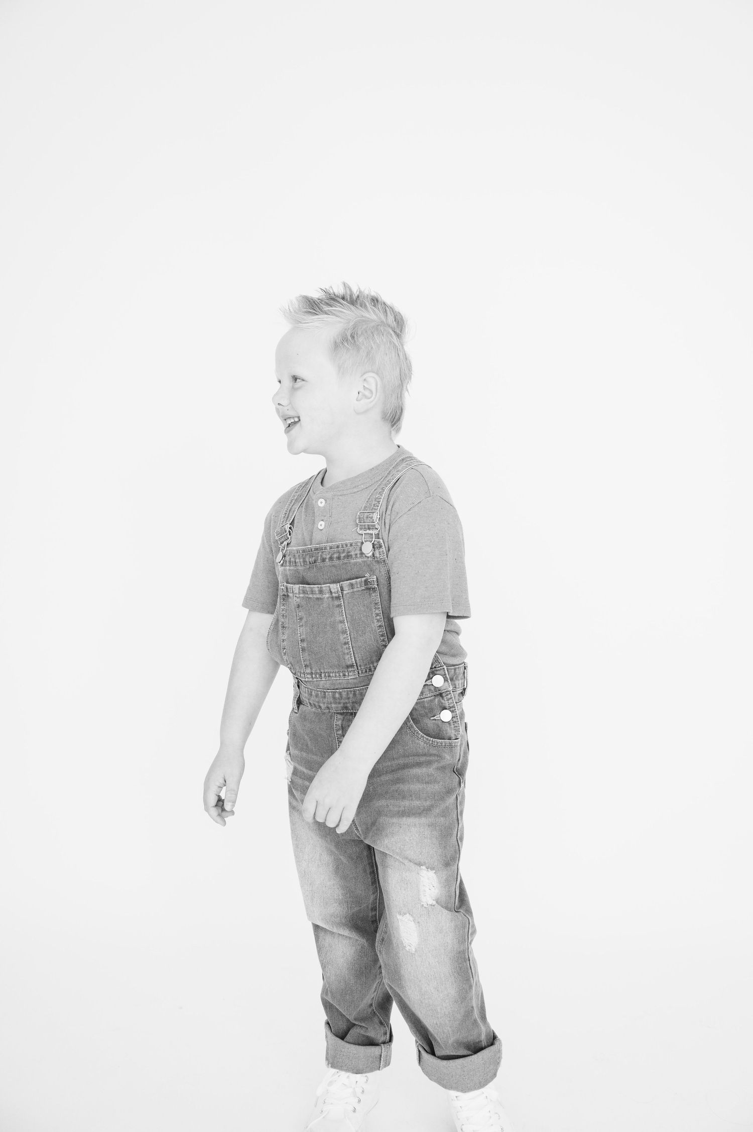 Child wearing overalls and t-shirt in black and white portrait against white background.
