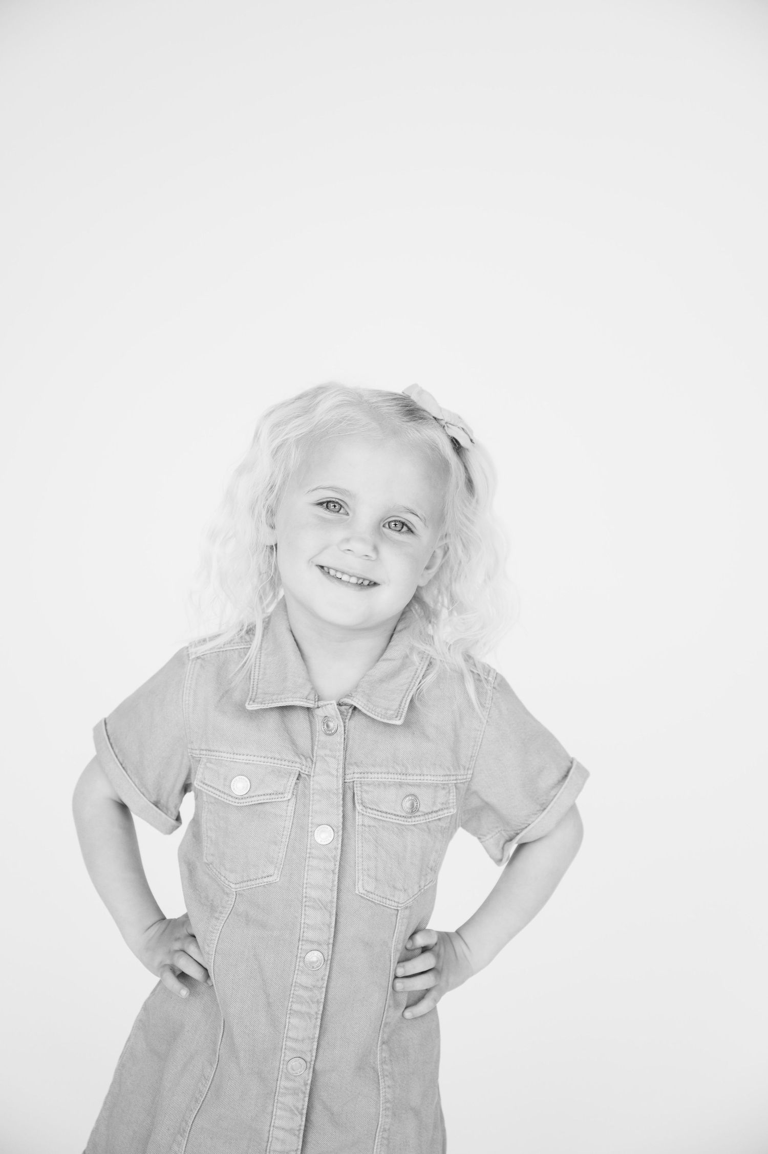 Black and white photo of a girl in denim dress posing with hands on hips against white backdrop.