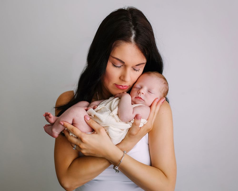 A tender moment shows a woman in a white sleeveless top cradling a sleeping newborn baby against a gray background.