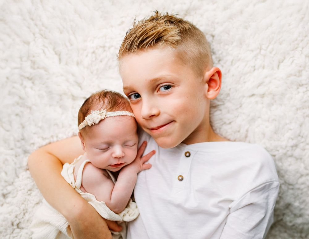 A young boy in a white button-up shirt gently holds a sleeping baby with a delicate headband on white fabric.