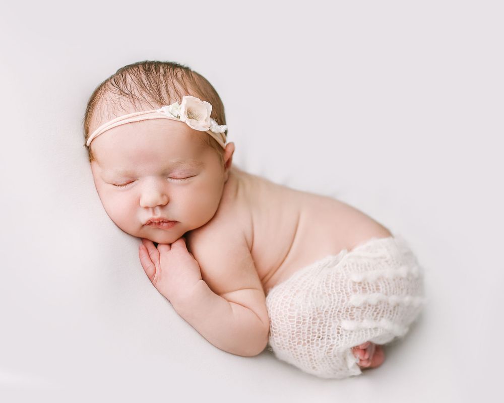 Peaceful sleeping newborn wearing white knit diaper cover and delicate bow headband on light background.