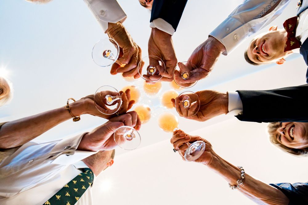 A circle of hands holding champagne glasses together in a celebratory toast against a bright background.