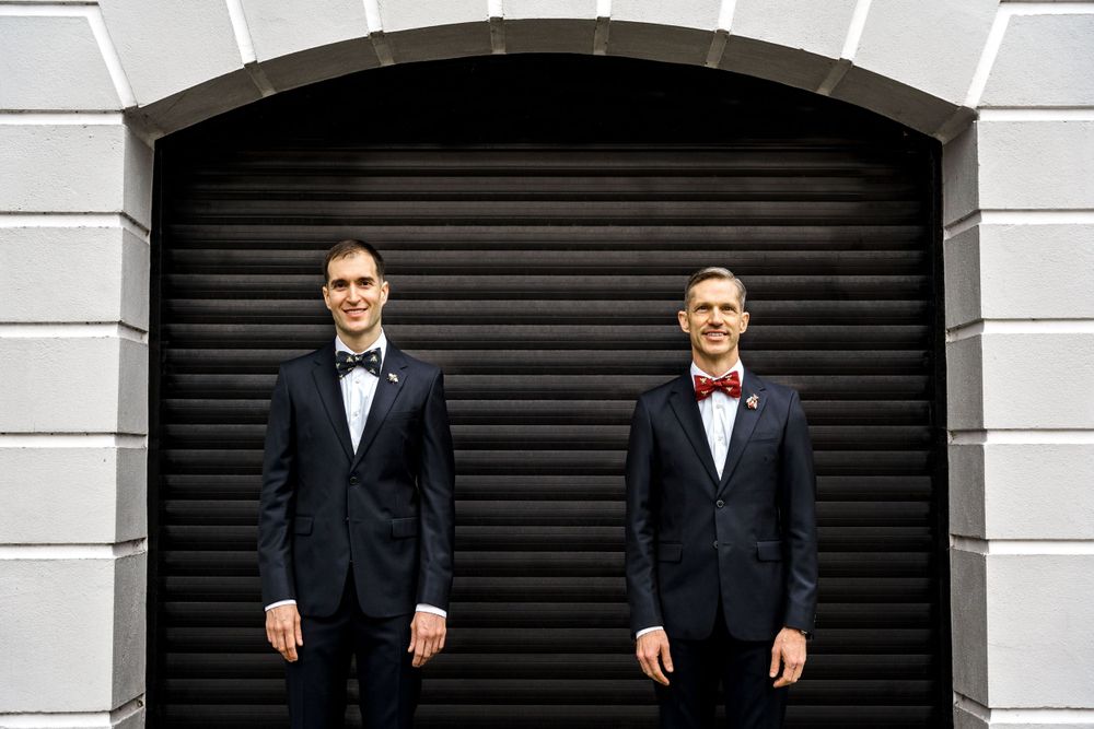 Two people in matching black tuxedos and bow ties stand in front of a dark garage door.