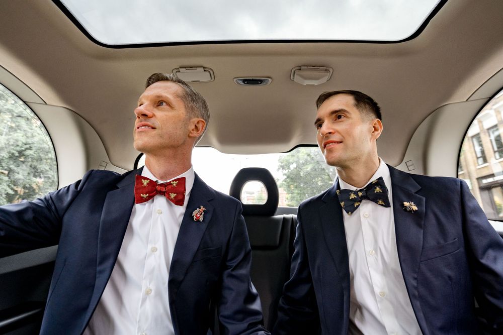 Two people wearing navy suits and bow ties sit in the backseat of a car with a sunroof.