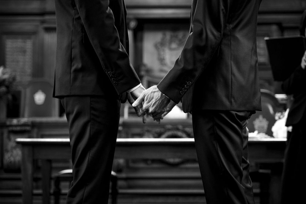 Black and white image of two people holding hands while wearing formal suits.