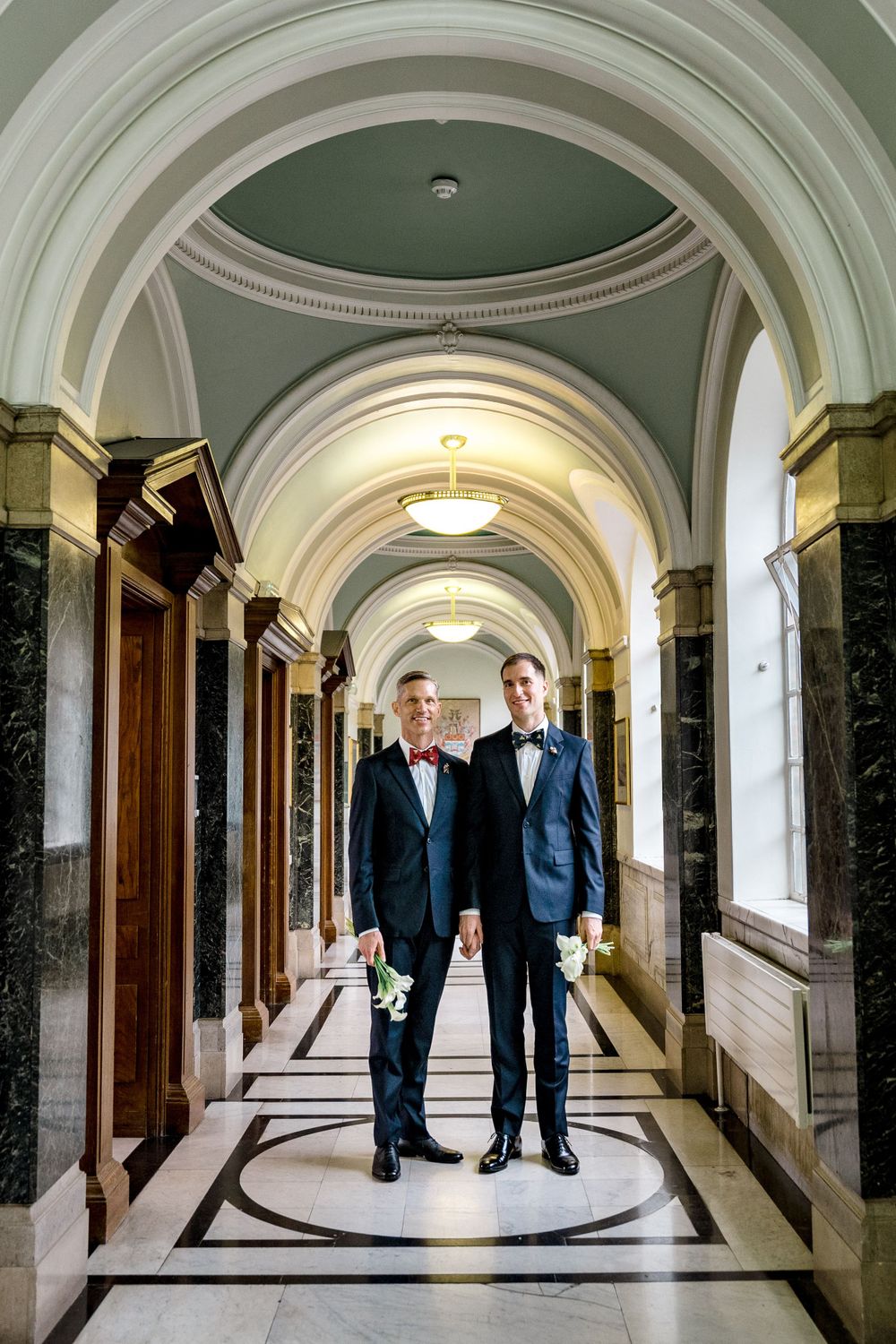 Two people in formal attire standing in an elegant hallway with arched ceilings and marble columns.