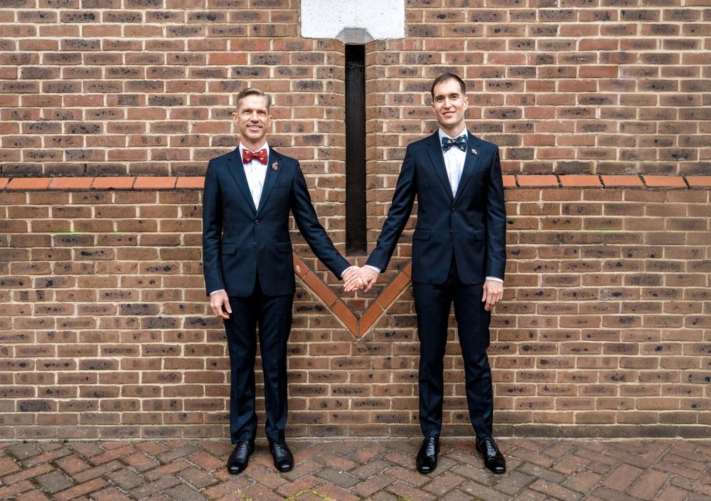 Two people in matching tuxedos holding hands against a brick wall with a basketball hoop.