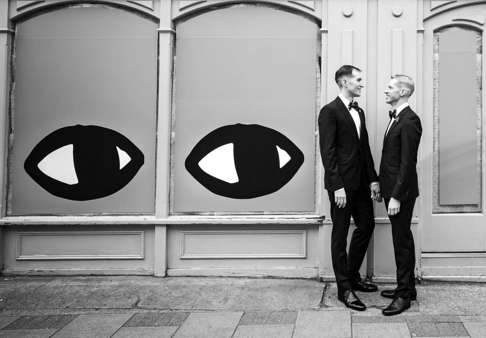 Two people in formal wear standing between large decorative eye murals on a building exterior.
