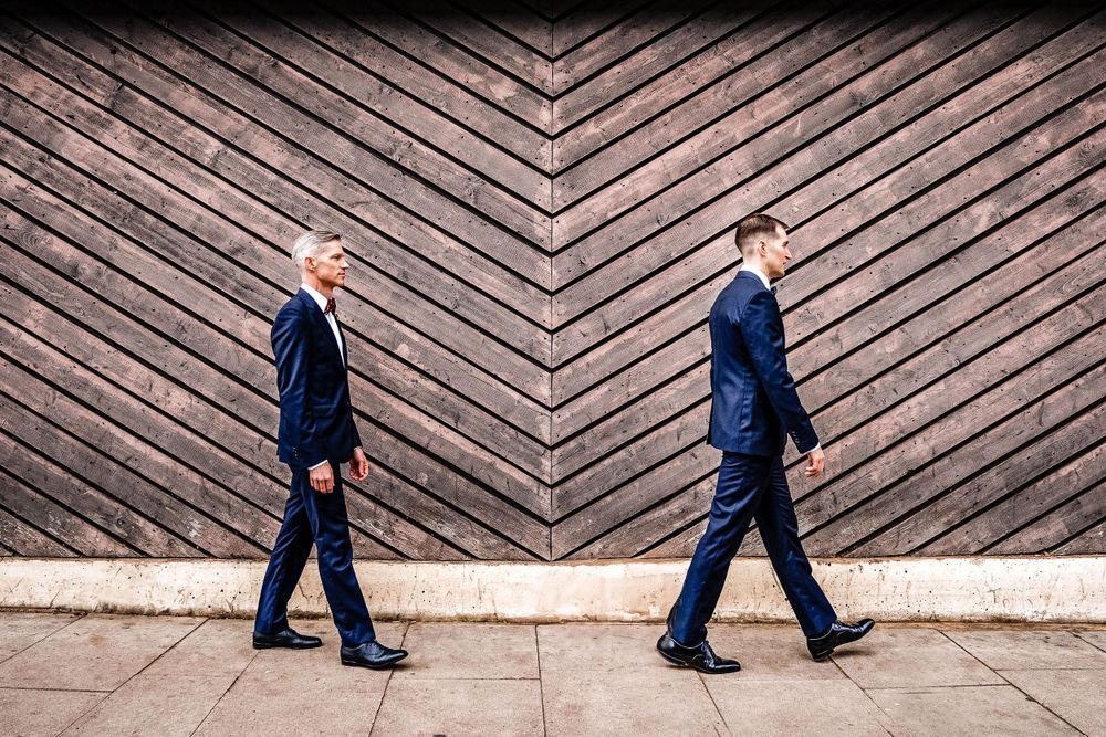 Two people in suits walking in opposite directions against a geometric wooden wall pattern.