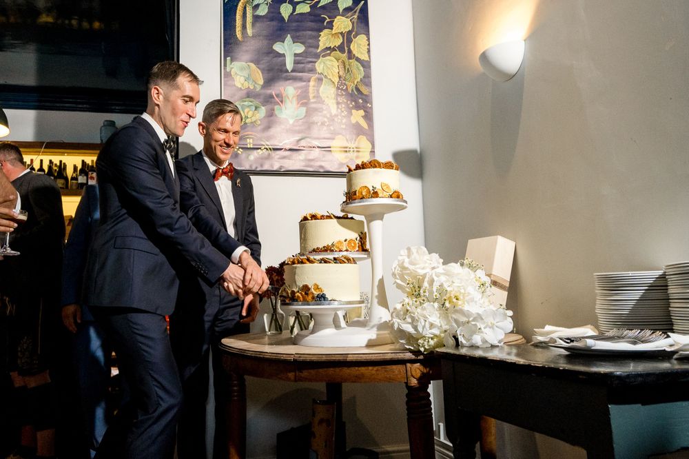 Two people in dark suits cutting their wedding cake together at a reception.
