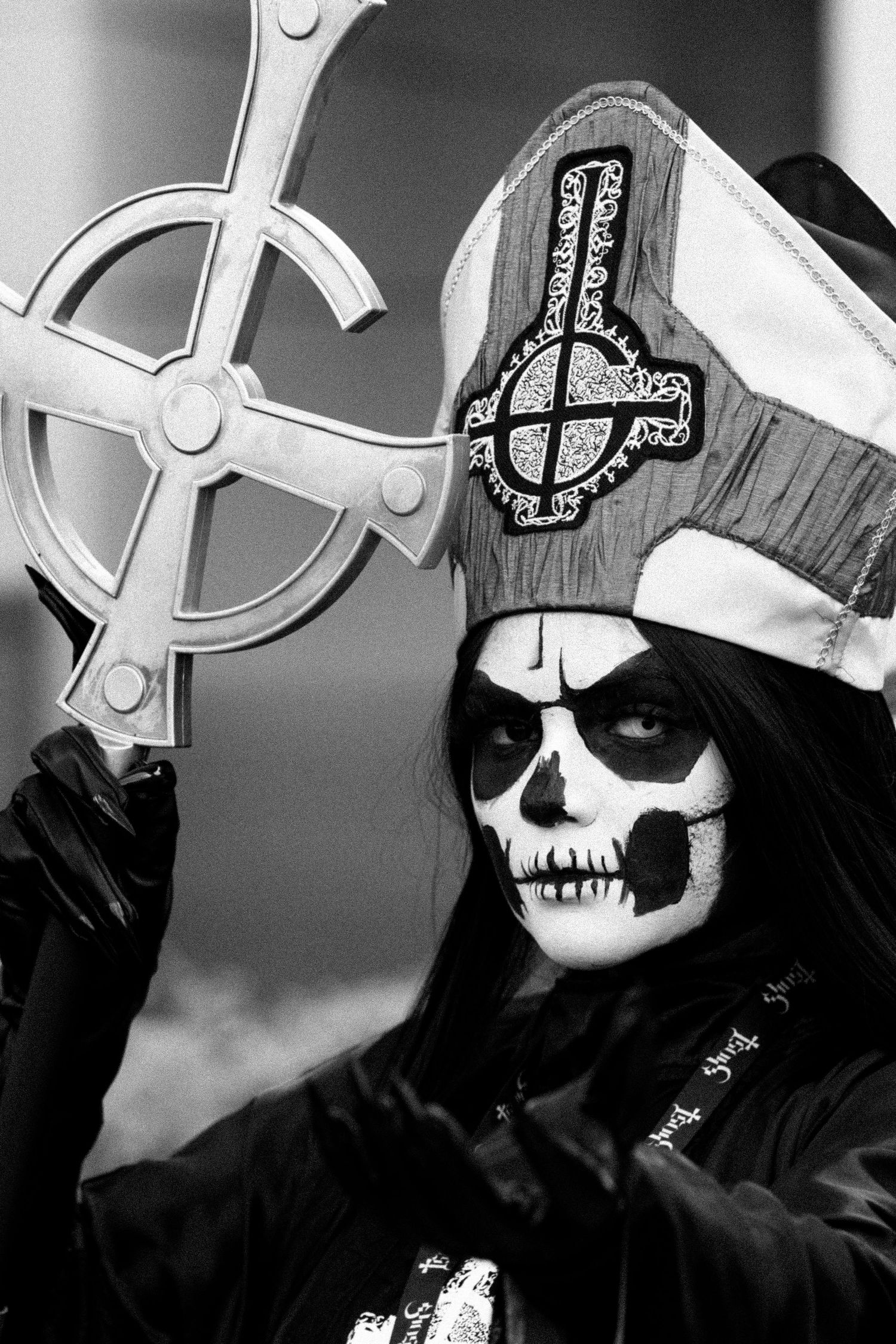Person in black cloak wearing skull face paint and religious headpiece holds up an inverted cross in dark setting. | Shot By Lo™