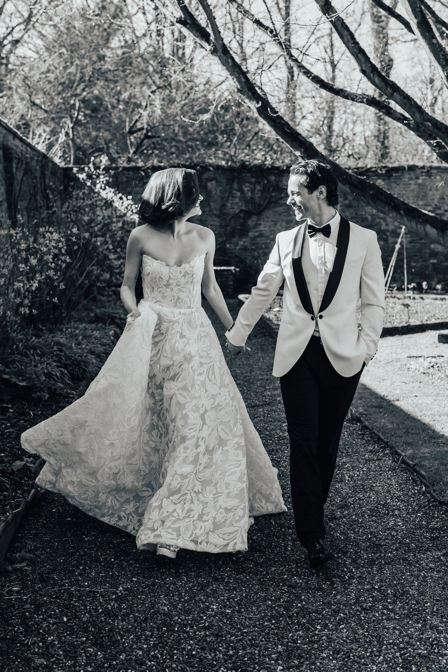 Black and white photo of a bride and groom holding hands, walking together outdoors in elegant wedding attire.