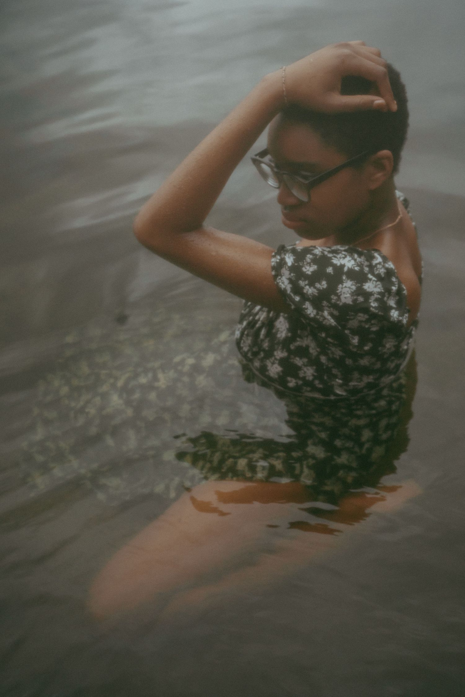 A person in a floral swimsuit and glasses wades in dark water with an artistic blurred effect.