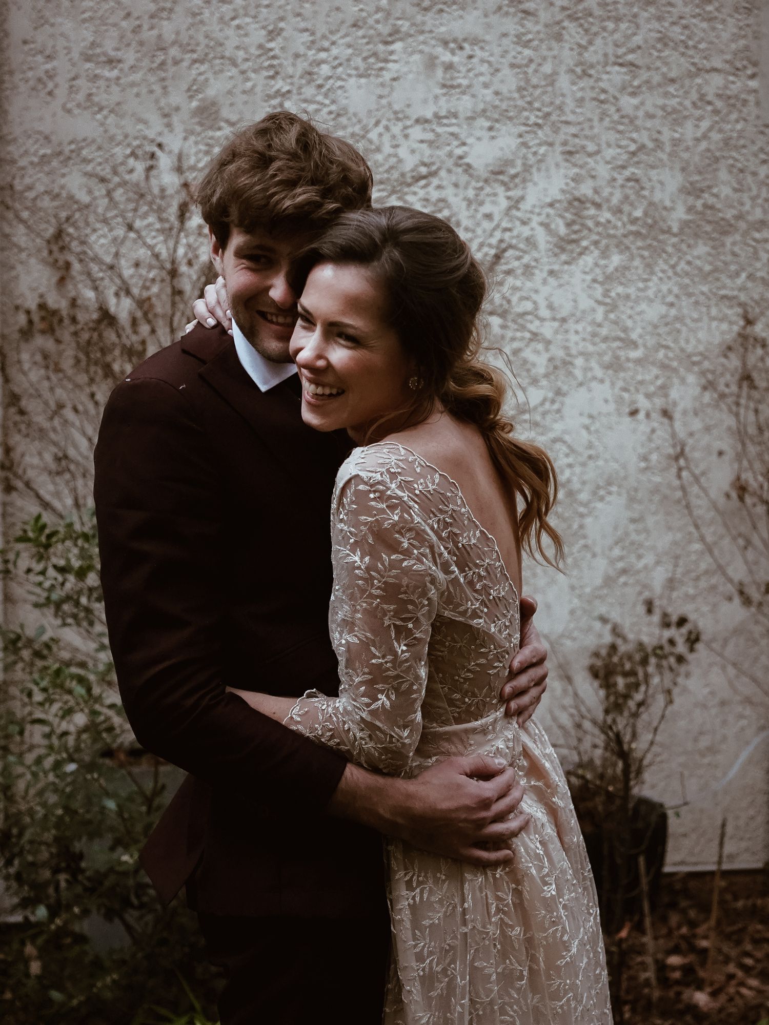 Groom embracing bride from behind outdoors, bride smiling in a beautiful lace backless wedding gown.