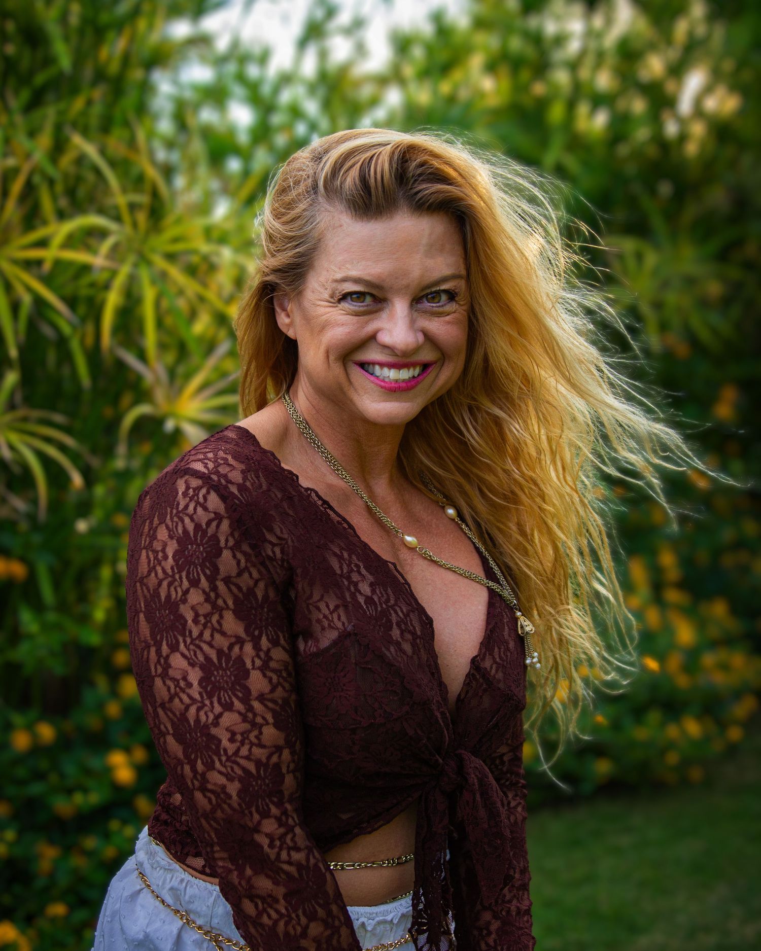 A portrait of someone in a burgundy lace top with flowing blonde hair against greenery.