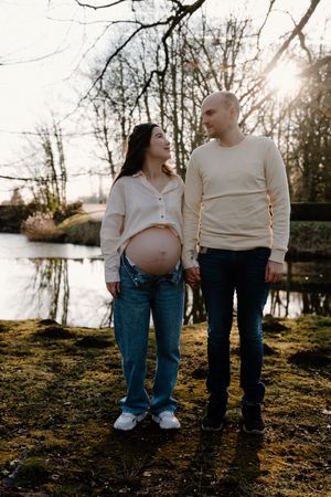 Expecting couple in casual denim and white tops pose near a lake at golden hour.