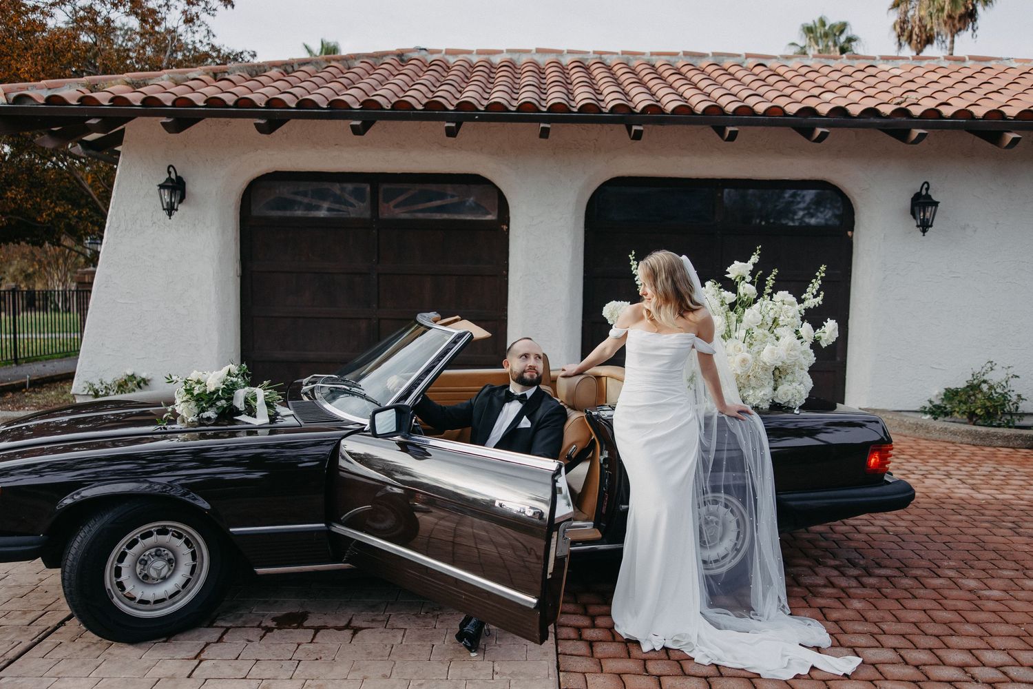 Wedding couple with vintage black convertible outside Spanish-style garage with terracotta roof.