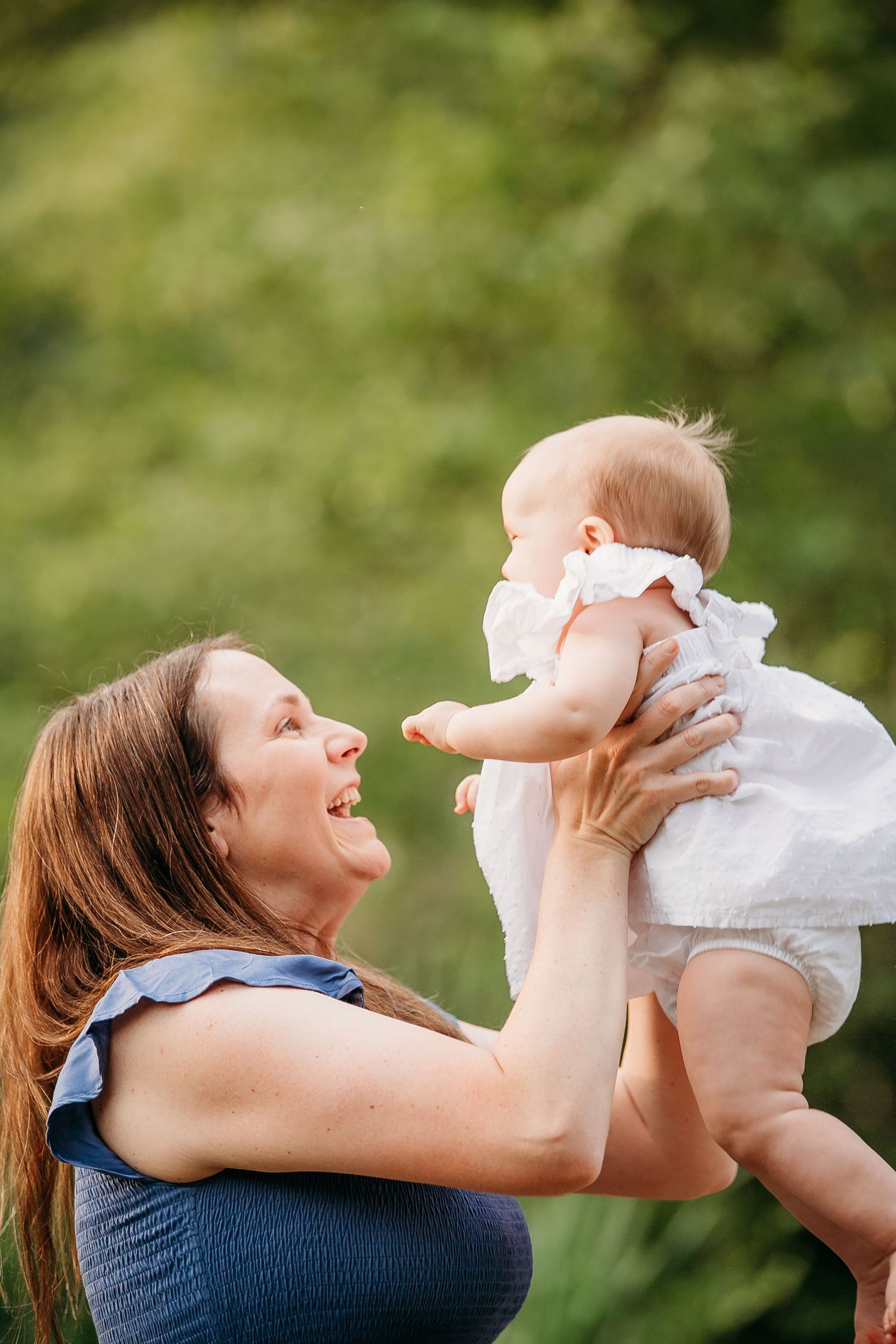 Joyful mother lifts smiling baby in white dress outdoors with lush green background.