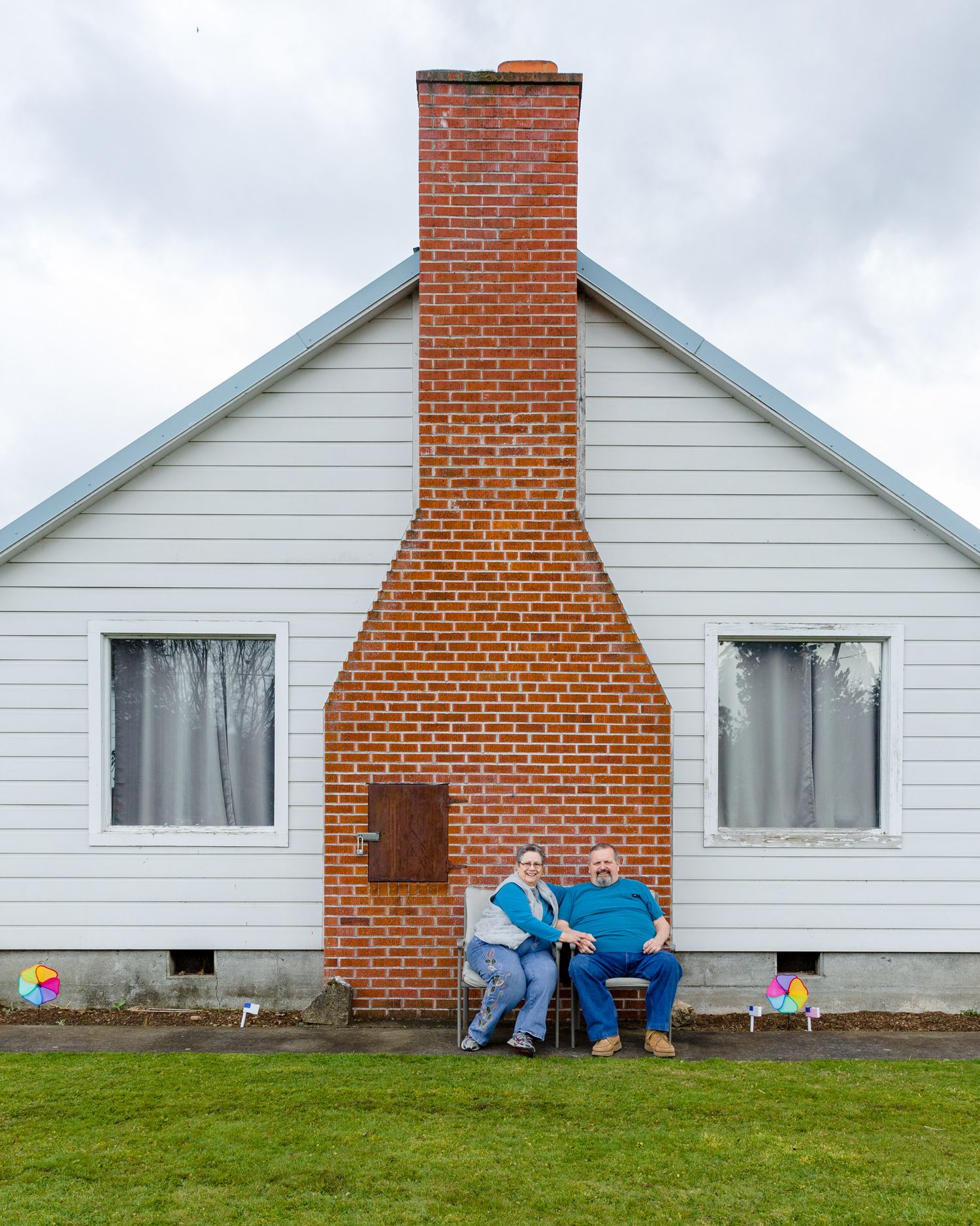 A white house with a prominent brick chimney and two people sitting next to it on a grassy lawn.